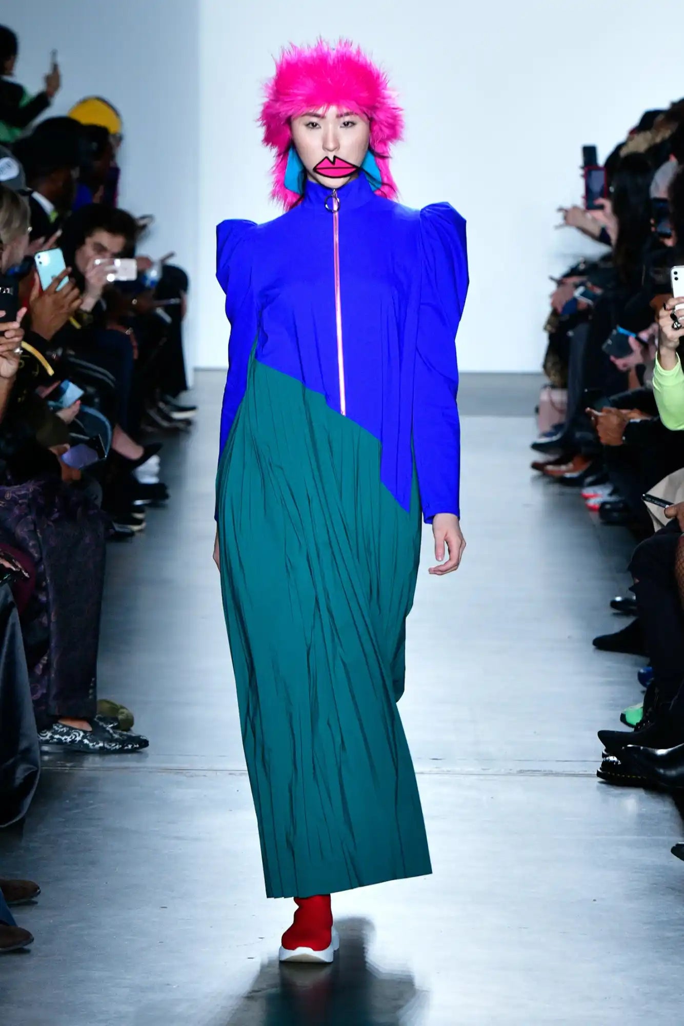 Bold color-blocked outfit featuring a royal blue top with structured shoulders, emerald green skirt, bright pink wig, and red shoes on a fashion runway.