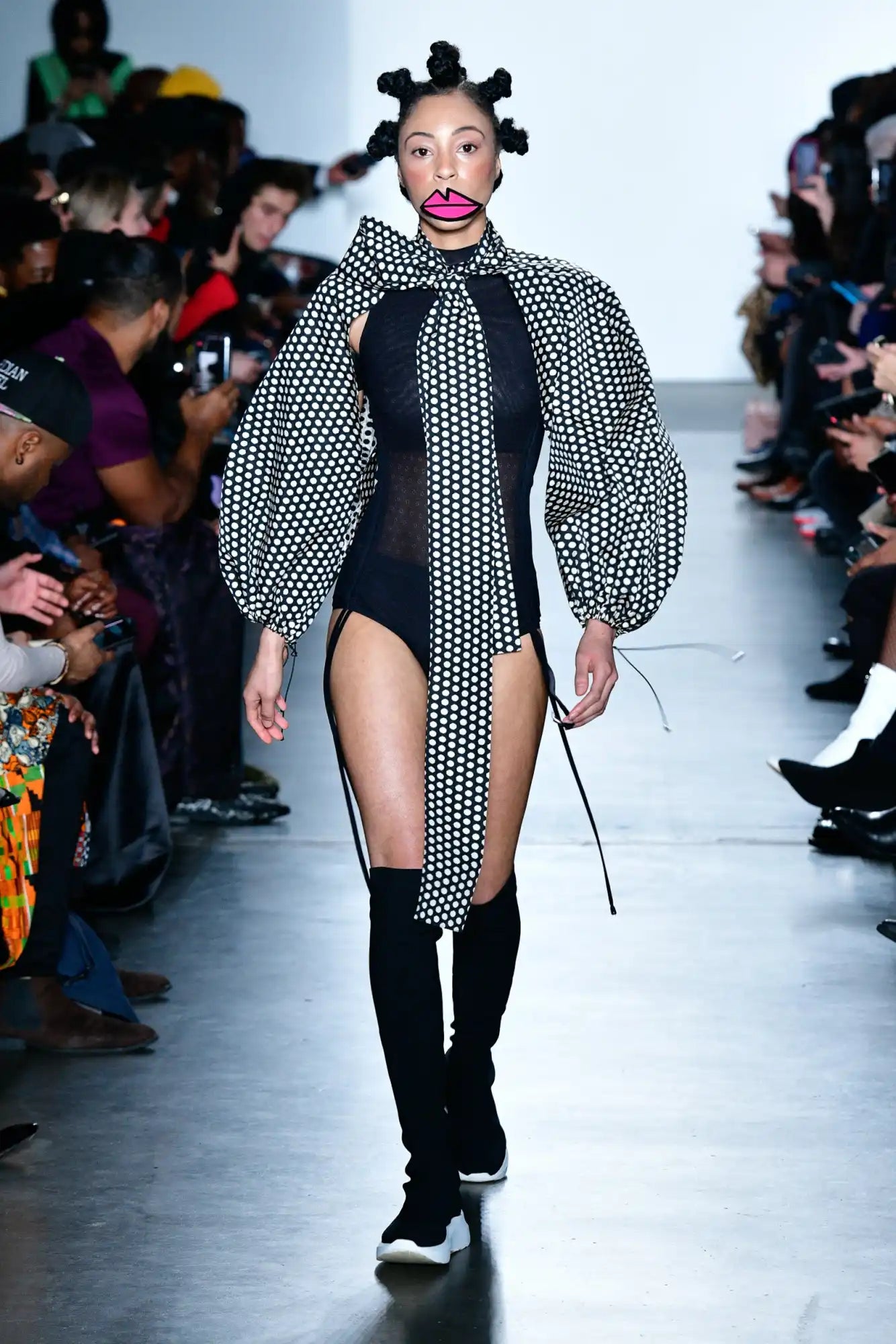 A fashion model wearing a black bodysuit with a dramatic black-and-white polka dot scarf and knee-high boots on a runway.