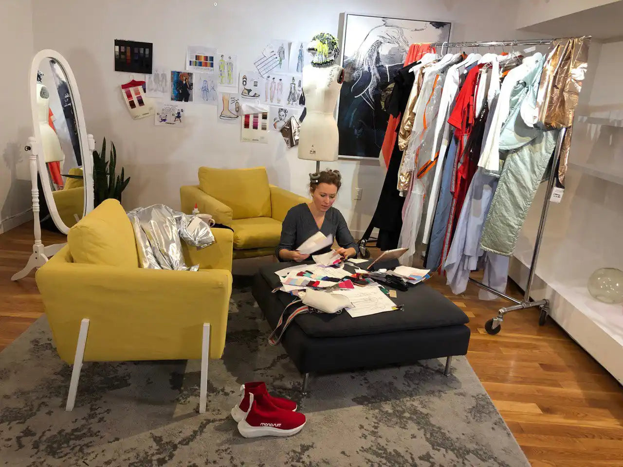 A person sitting on a black ottoman surrounded by clothing and yellow chairs.