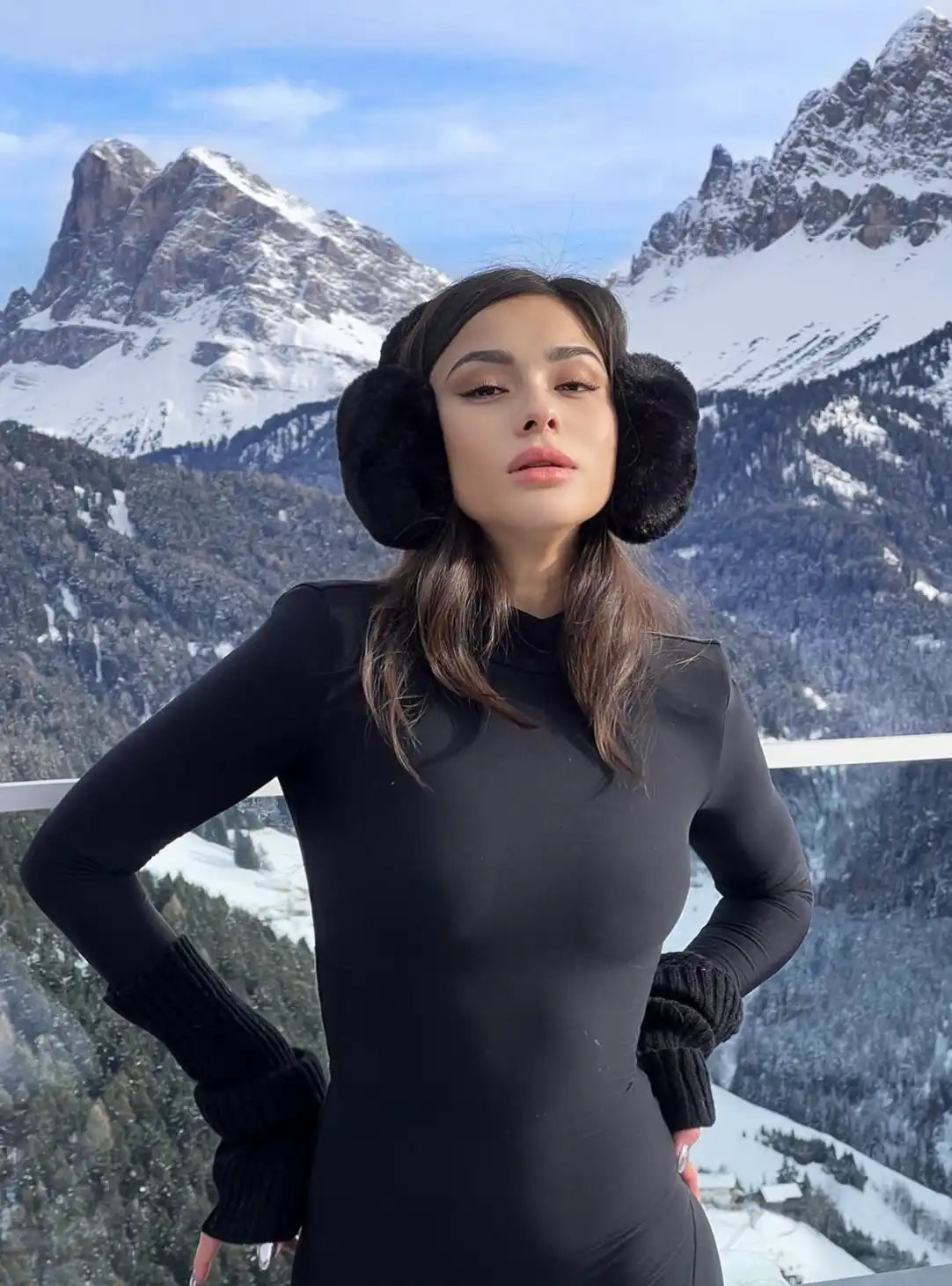 A person wearing a black turtleneck and winter earmuffs against snowy mountains.