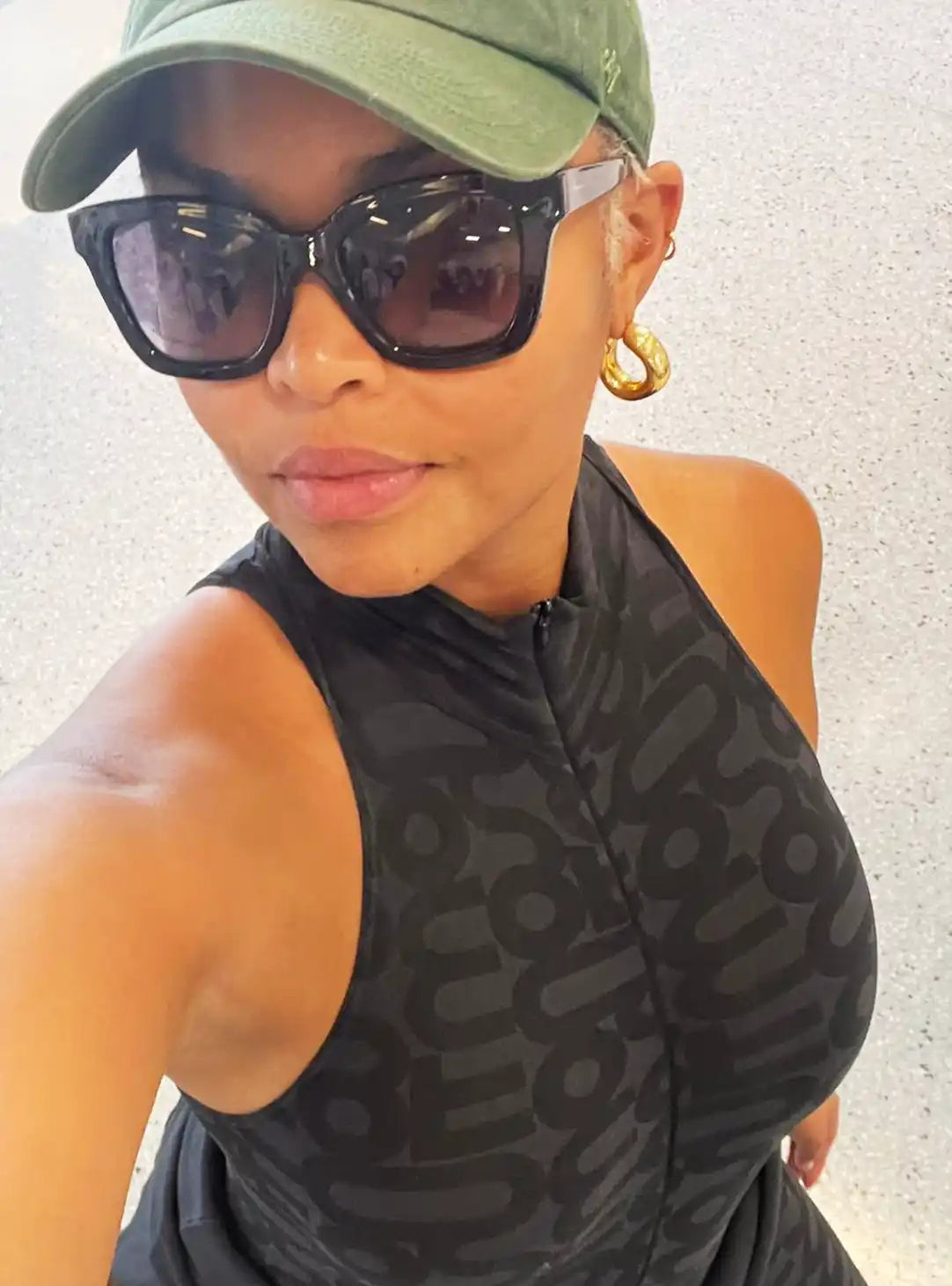 A selfie showing someone wearing a green baseball cap, dark sunglasses, and a sleeveless black patterned top.